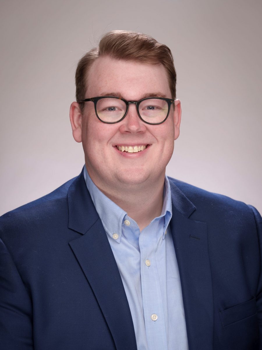 Image of Isaac smiling, with a navy blue suit jacket and light blue collared-shirt